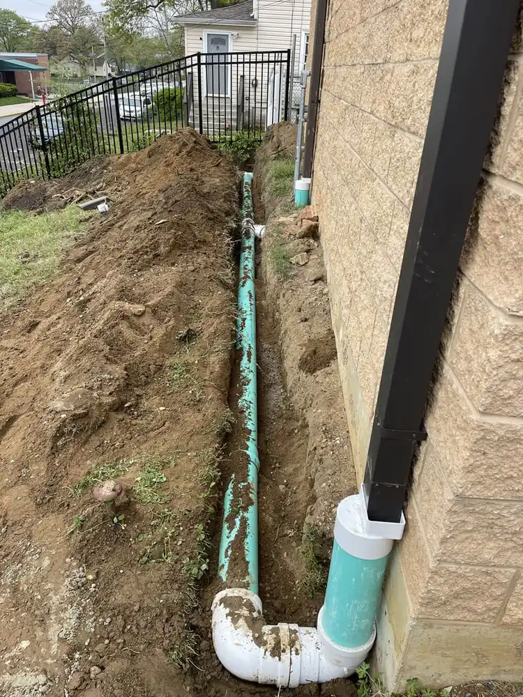 How Much Does Hydro Jetting Cost? White and green drain pipe running along a house foundation and connecting into a downspout