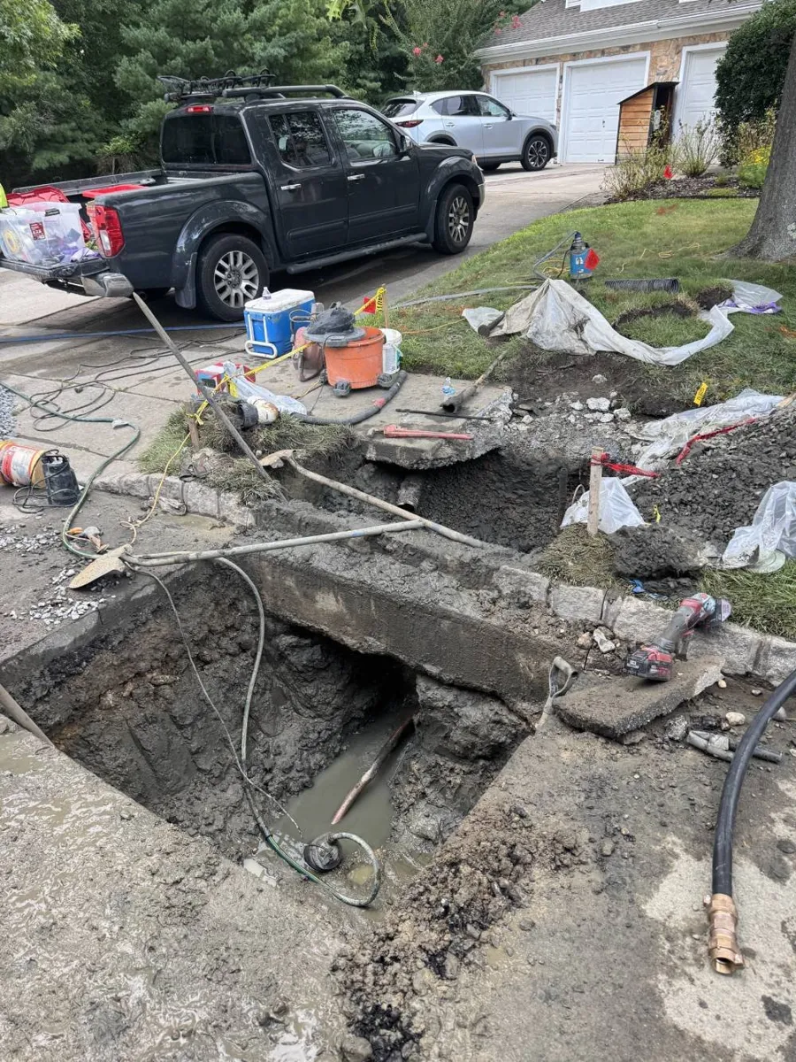How Much Does Trenchless Sewer Line Replacement Cost? Open access pit beside a driveway with a pickup truck and tools staged on the lawn