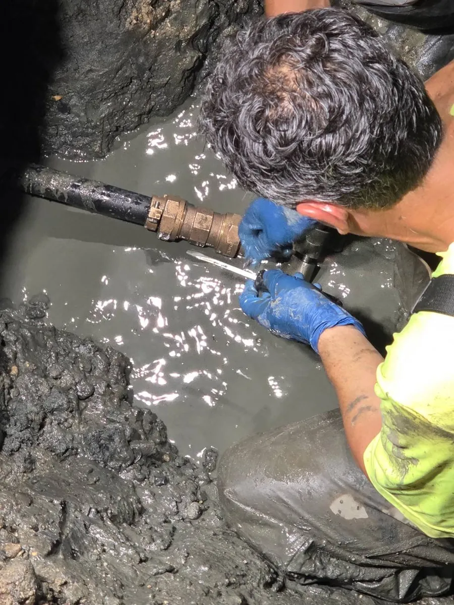Signs Your Water Line Needs to Be Replaced Technician in gloves working on a metal pipe fitting in a water filled excavation